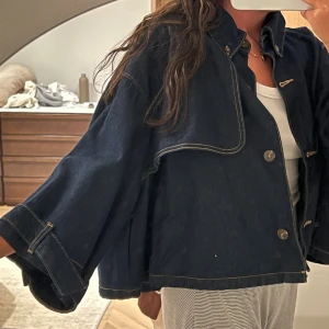 Oversized jeansjacka från Zara - Mörkblå oversized jeansjacka med poncho känsla från Zara med vida ärmar och snygga kontrastsömmar. Jackan har knappar framtill, hög krage och cool detalj med asymmetrisk panel på bröstet. Perfekt för dig som gillar en avslappnad och trendig look.