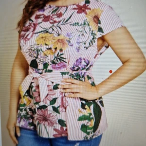 Blommig randig blus med knytband 3XL - Superfin blus i 3XL med rosa och vita ränder, täckt av färgglada blommor i gult, lila, grönt och vinrött. Modellen har korta ärmar, rund hals och ett snyggt knytband i midjan som ger en markerad siluett.  Bystmått 130-138. Ingen strech I tyget. 