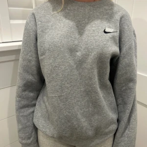 Grå sweatshirt från Nike - En klassisk grå sweatshirt från Nike med broderad logga på bröstet. Tröjan har rund halsringning och långa ärmar, tillverkad i mjukt material som är perfekt för chill dagar eller träning. Enkel och stilren design som funkar till det mesta.