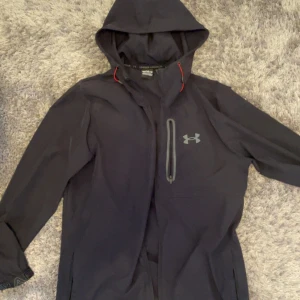Svart vindjacka från Under Armour - Snygg svart vindjacka från Under Armour med huva och grå detaljer. Jackan har en vertikal bröstficka med dragkedja och logga på framsidan. Perfekt för blåsiga dagar och sportiga aktiviteter. Lätt och smidig design med röda inslag vid halsen.