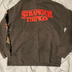 Stranger Things svart tröja från Zara - Mörk grå tröja från Zara med stort rött Stranger Things-tryck på bröstet, tecknade figurer från serien på ena ärmen och en stor Demogorgon-illustration på ryggen. Mjuk bomullskvalitet och ribbade muddar vid ärmslut och nederkant. Perfekt för fans av serien!