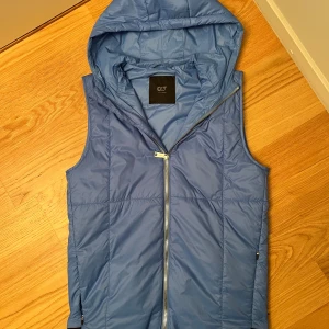 Blå dunväst med huva från AlphaTauri - Snygg blå dunväst från AlphaTauri med huva och hel dragkedja framtill. Västen har två fickor med dragkedja och en svart kant nertill med logotyp. Perfekt för lager-på-lager och chill stil. Storlek S-M