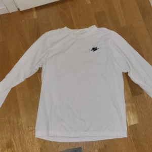 Vit långärmad Nike tröja - Säljer en stilren vit långärmad tröja från Nike med klassisk svart logga på bröstet. Perfekt till chill dagar eller när du vill ha en enkel och clean look. Skön passform och mjukt material.