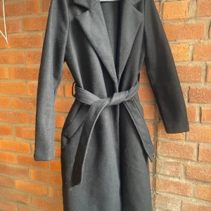 Svart kappa från Vero Moda XS - Stilren svart kappa från Vero Moda i storlek XS. Jackan har klassisk krage, knytskärp i midjan och långa ärmar. Perfekt för dig som gillar en enkel och tidlös look. Materialet är mjukt och känns skönt mot kroppen.