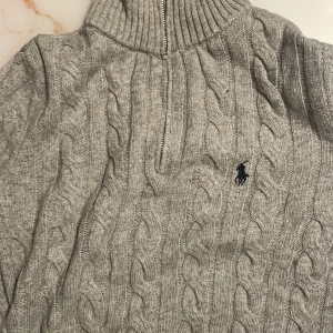Grå stickad half zip-tröja Polo Ralph Lauren - Kabelstickad grå tröja från Polo Ralph Lauren med hög krage och half zip-dragkedja med läderdetalj. Klassisk broderad logga på bröstet. Perfekt för lager-på-lager och chill höst- eller vinterstil.