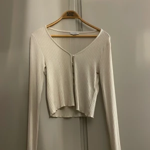 Vit ribbad topp med pärlknappar Amisu - Säljer en vit ribbad topp från Amisu i storlek S. Toppen har lång ärm, croppad passform och v-ringning. Stängs med dekorativa pärlknappar framtill. Perfekt att styla med jeans eller kjol för en trendig look.