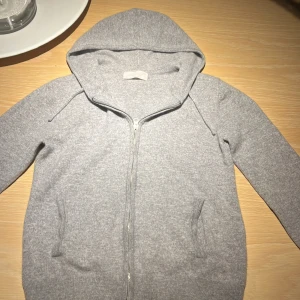 Grå Kashmir hoodie med zip - Grå hoodie med dragkedja framtill och huva med snören. Tröjan har två fickor på sidorna och är långärmad med ribbade muddar. Nypris är ca 1000kr
