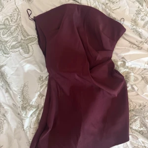 Vinröd axelbandslös topp - Säljer en vinröd axelbandslös topp med figurnära passform. Toppen har en enkel och stilren design med lätt drapering framtill. Perfekt för dig som vill ha en elegant look med en djup färgton.