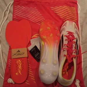 Adidas F50 Elite fotbollsskor i vitt och rött - Säljer ett par Adidas F50 Elite fotbollsskor i vitt med röda och svarta detaljer. Skorna har snörning, platt sula med dobbar och levereras med extra innersulor och en matchande röd påse. Perfekta för dig som vill sticka ut på planen!