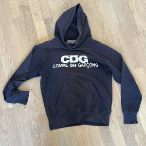 Mörkblå CDG hoodie Comme des Garçons - Snygg mörkblå hoodie från Comme des Garçons Good Design Shop i storlek S. Stor vit CDG-logga på bröstet, klassisk huva och magficka. Tillverkad i mjuk bomull med ribbade muddar och lång ärm. Perfekt för dig som gillar streetwear och vill sticka ut. Hoodien har tyvärr ett flertal slitningar, men som enligt mig gör det till en ännu mer vintage och unik look. Därav priset, inte hugget i sten men känns enligt mig relativt rimligt med tanke på nypris. Kom gärna med frågor! 
