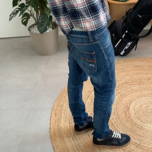 Blå selvedge Nudie Jeans med slitningar - Säljer ett par blå jeans från Nudie Jeans med orange sömmar och snygga slitningar på både fram- och bakficka. Jeansen har klassisk femficksdesign, normal passform och är tillverkade i mjukt bomullsjeans. Perfekta för dig som gillar en avslappnad och trendig look.