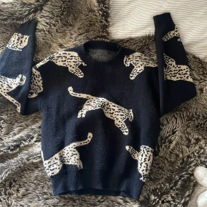 Svart leopardmönstrad stickad tröja Cider - Cool svart stickad tröja från Cider med beige leopardmönster över hela plagget. Tröjan har rund hals, långa ärmar och en loose passform som ger en avslappnad vibe. Perfekt för dig som vill sticka ut med ett djuriskt print.