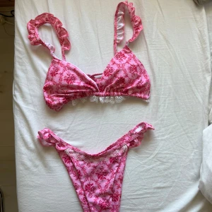 Rosa blommig bikini med spets och volang - Fin rosa bikini med blommigt mönster och volang på axelbanden och vid midjan/kanterna. Både överdel och underdel har vita spetsdetaljer. Aldrig använd!🩷🌸