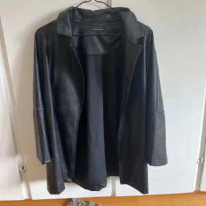 Svart skinnjacka från Black Colour - Svart skinnjacka från Black Colour i oversized modell. Jackan har en enkel krage, långa ärmar och medföljande bälte för att knyta i midjan. Snygg och stilren look som passar perfekt till lager-på-lager. Materialet är mjukt och ger en cool vibe. Aldrig använd. 