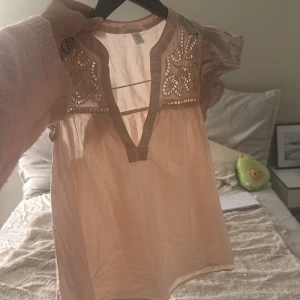 Ljusrosa blus med broderad detalj - Supersöt ljusrosa blus med v-ringning och broderade detaljer upptill. Blusen har korta, volangiga ärmar och är tillverkad i ett tunt, luftigt material som känns perfekt för varmare dagar. Den har en romantisk och bohemisk vibe med sina fina utskärningar och lätta tyg.