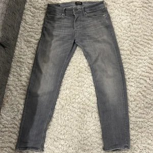 Grå jeans från Jack&Jones  - Snygga grå jeans från Nudie med klassisk femficksdesign och diskreta slitningar. Jeansen har smal passform och raka ben, tillverkade i mjuk denim med stretch för extra komfort. Perfekta för dig som gillar en stilren och modern look.