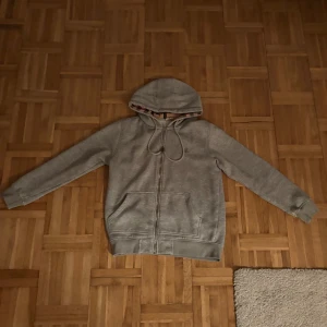 Grå Burberry hoodie med dragkedja - Grå hoodie från Burberry med klassiskt rutigt foder i huvan och broderad logga på bröstet. Tröjan har dragkedja framtill, snörning i huvan och två fickor. Tillverkad i mjuk bomullsmix, perfekt för kyliga dagar. Minst 700