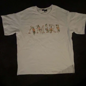 Vit blommig t-shirt från Amiri - Vit t-shirt från Amiri med blommigt tryck över loggan på bröstet. Klassisk passform och rund halsringning. Perfekt för dig som vill ha en stilren men ändå unik look. Materialet är mjuk bomull som känns skönt mot huden.