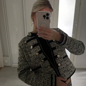 Svart och guld kavaj från Flash Woman - Säljer en svart kavaj med guldigt brokadmönster från Flash Woman. Jackan har dekorativa guldknappar, svarta detaljer och en rak, kortare passform. Perfekt för dig som vill sticka ut med en unik och elegant look.