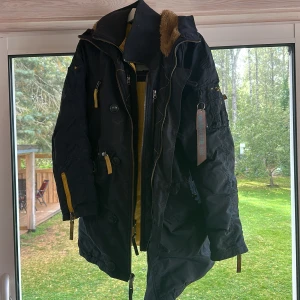 Svart parkajacka Alpha Industries S - Säljer en svart parkajacka från Alpha Industries i storlek S. Jackan har gula detaljer, huva med fuskpäls, flera dragkedjor och knappar samt patch på ärmen. Fodret är gult och jackan har en längre modell med snygga detaljer och praktiska fickor.