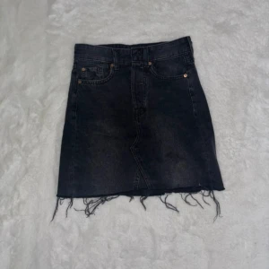 Svart jeanskjol med fransig kant - Snygg svart jeanskjol med rå, fransig nederkant och klassiska fickor fram. Kjolen har en rak passform och stängs med knapp.