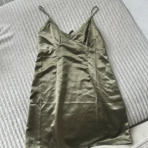 Olivgrön satinklänning med spets - Olivgrön kort klänning i satin med tunna axelband och spetsdetaljer vid urringningen. Klänningen har snörning i ryggen för en snygg passform och glansigt tyg