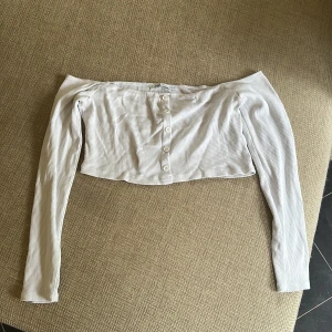 Vit ribbad offshoulder topp från Zara - Säljer en vit croppad offshoulder topp från Zara med långa ärmar. Toppen är ribbad och har knappar framtill för en snygg detalj. Perfekt för dig som gillar trendiga och stilrena plagg med en twist.