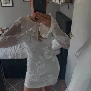Vit blommig kortklänning med öppen rygg - Supersnygg vit kortklänning med långa transparenta ärmar och stora blommiga broderier. Klänningen har en öppen rygg och utsvängda ärmslut som ger en extra  vibe. Perfekt studentklänning!! 