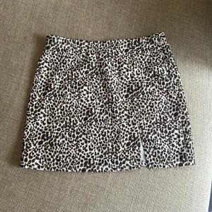 Leopardmönstrad kortkjol med slits - Snygg kortkjol med leopardmönster i svart och brunt på beige botten. Kjolen har en liten slits framtill som ger en extra detalj. Perfekt för dig som vill sticka ut med ett djurmönster. Materialet känns mjukt och följsamt, troligtvis bomull eller polyester.