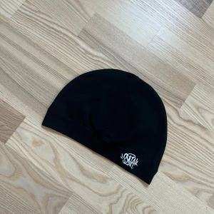 Svart mössa från syna  - Svart, stilren mössa från syna med broderad vit logga framtill. Mjuk och stretchig modell i slät bomullsmix som sitter skönt på huvudet. Perfekt för kalla dagar och enkel att matcha med olika outfits. Oanvänd