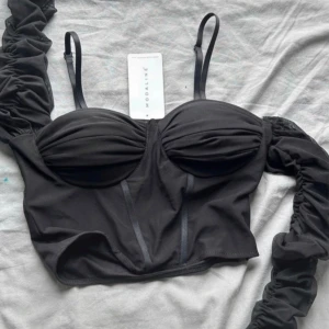 Svart korsettopp från Modaline oanvänd  - Trendig svart korsettopp från Modaline med avtagbara meshärmar och smala axelband. Toppen har vadderade kupor med rynkade detaljer och markerade sömmar framtill. Sitter croppad och har en snygg, figurnära passform. Perfekt för att lyfta vilken outfit som helst.