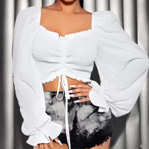 Vit croppad blus med puffärmar - Säljer en vit croppad blus med långa puffärmar och smockad, ribbad kropp. Toppen har en söt volangkant och snörning framtill som ger en snygg detalj. Materialet är lätt och krispigt, perfekt för dig som gillar romantisk stil med modern twist.