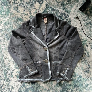 Grå jeansjacka med fransade kanter - Snygg grå jeansjacka med fransade kanter och markerade sömmar. Jackan har två stora knappar framtill, fickor med lock och coola detaljer längs krage och ärmslut. Figursydd passform och klassisk kavajkrage ger en unik look.