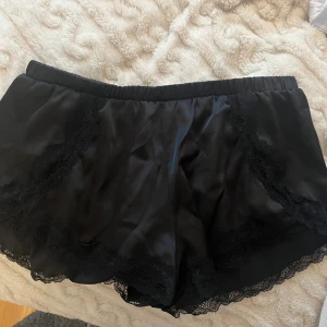 Svarta silkesliknande shorts med spets - Säljer ett par svarta shorts i silkesliknande material med bred resår i midjan. De har fina spetsdetaljer längs kanterna och en mjuk, flowy passform. Perfekta för en chill look eller som pyjamasshorts.