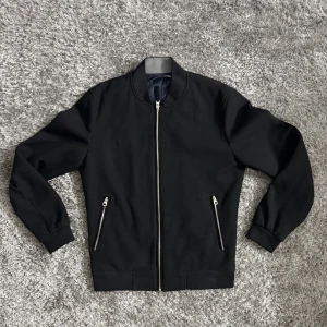 Svart bomberjacka från Zara Man - Stilren svart bomberjacka från Zara Man med klassisk rund krage och silverfärgade dragkedjor. Jackan har två snedställda fickor med dragkedja och är helfodrad i mörkblått tyg. Perfekt för dig som gillar en clean och modern look. Står M men passar S också 