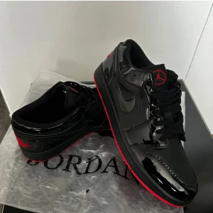 Nike Air Jordan 1 Low svart/röd 43 - Säljer ett par Nike Air Jordan 1 Low i svart och rött. Skorna har glansiga detaljer i lack, klassisk röd yttersula och svarta snören. Röd Jumpman-logga på plösen och Air Jordan Wings på hälen. Perfekta för dig som gillar streetwear och sneakers med attityd.