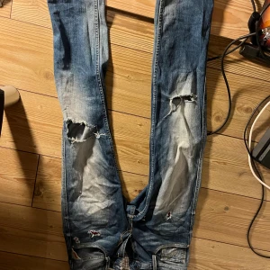 Replay jeans med slitningar och hål - Säljer ett par blå Replay jeans med coola slitningar och hål på knäna. Jeansen har klassisk femficksdesign, tydliga slitdetaljer och kontrastsömmar. Märkespatch bak i midjan och röd Replay-detalj vid fickan. Perfekta för dig som gillar en avslappnad och edgy stil. 