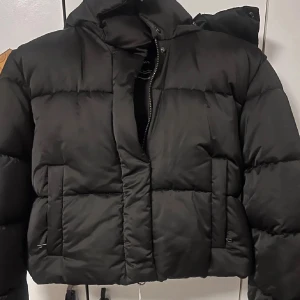 Svart pufferjacka med dragkedja - Säljer en svart pufferjacka med hög krage och dragkedja framtill. Jackan är quiltad och har två stora fickor på sidorna. Perfekt för kalla dagar och ger en cool, oversized vibe. Materialet är syntet och jackan har långa ärmar med resår vid handlederna.