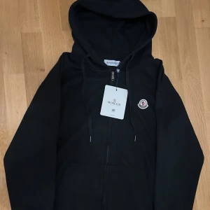 Moncler zip hoodie - Svart zip hoodie från Moncler med logga på bröstet, hel dragkedja framtill och luva. Hoodien är i storlek M och är i nyskick. Tveka inte på att höra av dig vid frågor.