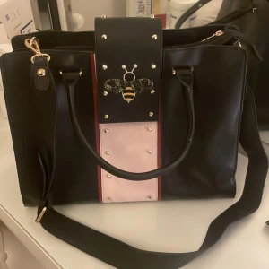 Svart handväska från Mary Kay  - Stilren svart handväska från Mary Kay med rosa och röda detaljer framtill, dekorerad med en cool bi i metall och pärlor. Väskan har både handtag och axelrem, guldfärgade nitar och dragkedja upptill. 