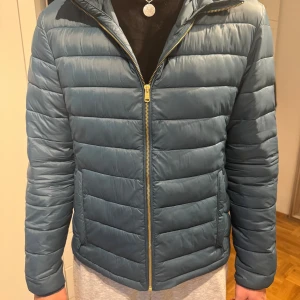 Blå pufferjacka med huva - Säljer en stilren blå pufferjacka med huva och guldfärgad dragkedja. Jackan har två fickor framtill och en klassisk passform. Perfekt för kyliga dagar och enkel att matcha med olika outfits. Kan gå ner i pris vid snabb affär!