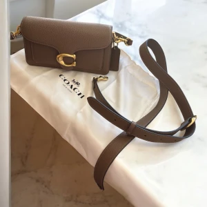 Brun axelväska i skinn från Coach - Snygg brun axelväska från Coach i äkta skinn med justerbar rem och guldfärgade metalldetaljer. Klassisk design med präglad logga och stort C-spänne framtill. Perfekt storlek för det viktigaste och stilren look.