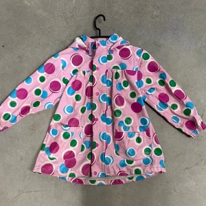 Rosa regnjacka med färgglada prickar - Snygg rosa regnjacka med stora färgglada prickar i blått, grönt och cerise. Jackan har huva, två stora fickor framtill och dragkedja. Perfekt för regniga dagar och sticker verkligen ut med sitt lekfulla mönster.