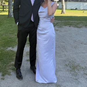 Almeria klänning från Adoore - Supersnygg ljuslila långklänning från Adoore i strl 36 med asymmetrisk axel och smala axelband. Använd en gång och i mycket bra skick! Klänningen har en elegant drapering framtill och en slits på sidan. Klänningen har dock mindre märken på ryggen (se bild 5) på två ställen från att ha varit insydd i midjan. Om köparen också önskar att sy in den så kommer detta dock inte att synas💕