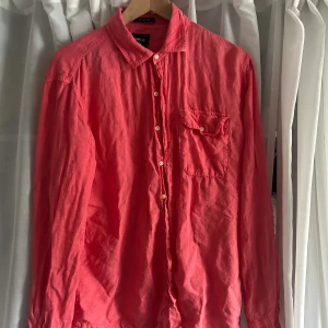 Rosa skjorta från Replay i bomull - Säljer en snygg rosa skjorta från Replay med klassisk krage, bröstficka med knapp och vita knappar framtill. Skjortan är långärmad och tillverkad i mjuk bomull, perfekt för en avslappnad men stilren look.