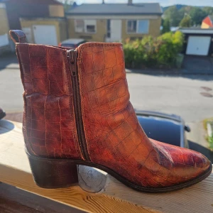 Bruna boots i krokopräglat skinn från Dasia - Säljer ett par bruna boots från Dasia i storlek 39. Skorna är i krokopräglat skinn med dragkedja på sidan och har en låg klack. Snyggt mönster och cool westernkänsla, perfekta för dig som vill sticka ut lite extra.