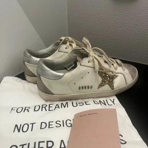 Golden Goose Superstar sneakers med glitterstjärna - Golden Goose Superstar sneakers i vitt skinn med grå mockadetaljer på tån och silverfärgad häl. Sidan pryds av en glittrig guldstjärna. Dom är köpa från plick för några tusen och där tillföljde denna påse + qr kod, som även tillföljer nu. Dom är i ett bra skick, men tyvärr har sulan därbak gått sönder lite på båda skorna. Det är inget som märks av men kan vara bra att nämna, samt lite skrap märken på skorna men det syns på bilderna (inge som syns mycket/märks av.  Skriv via frågor !! 💕💕💕💕