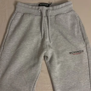 Grå mjukisbyxor från McKenzie - Snygga ljusgrå mjukisbyxor från McKenzie med resår och snörning i midjan. Byxorna har ribbade muddar vid bensluten och diskret logga på ena benet. Tillverkade i mjukt material som känns skönt mot huden. Perfekta för chill eller träning.
