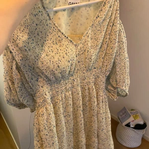 Blommig kortklänning från Ganni - Supersöt kortklänning från Ganni i ljusbeige med blått blommönster. Klänningen har v-ringning, puffiga korta ärmar och smockad midja som ger en snygg siluett. Tillverkad i ett lätt och luftigt material, perfekt för varma dagar. Tror att det är storlek M