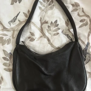 Svart axelväska från Gina Tricot - Stilren svart axelväska från Gina Tricot i mjukt skinn material. Väskan har en enkel, rundad form och stängs med en silvrig dragkedja. Perfekt storlek för det viktigaste och lätt att matcha till olika outfits.